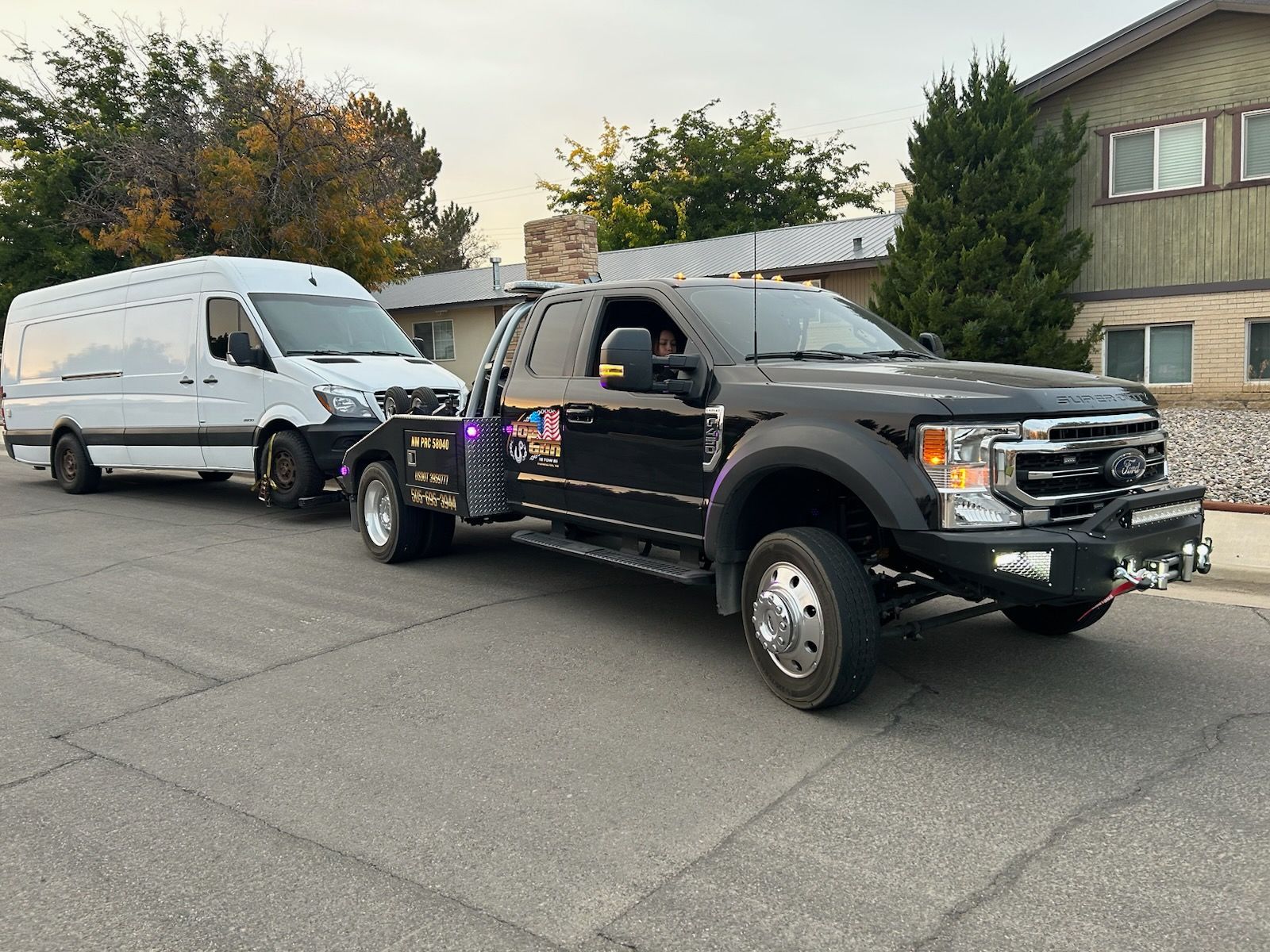 A black tow truck is towing a white van down a street.
