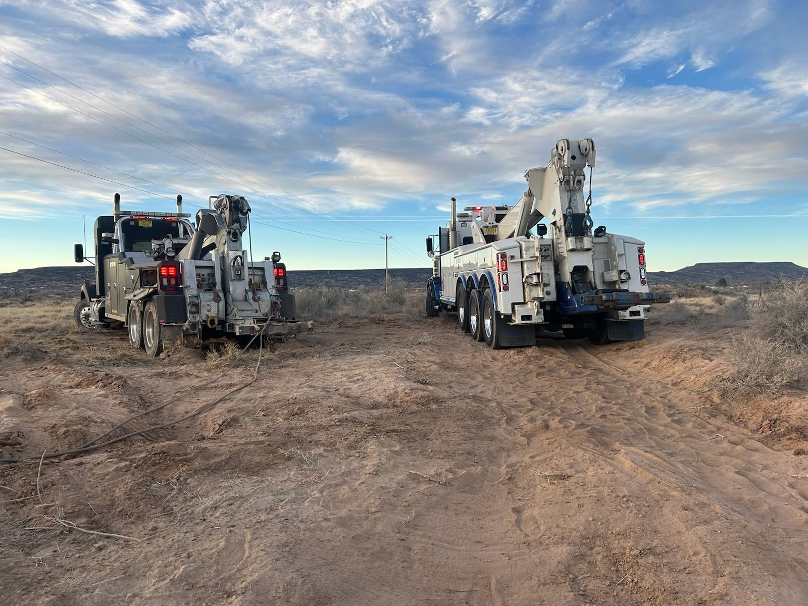 Two tow trucks are parked on the side of a dirt road.