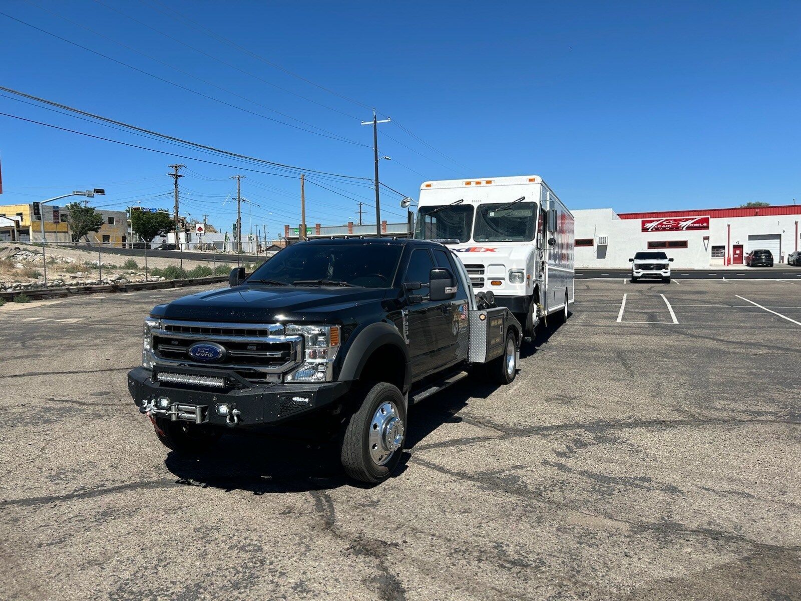 A black truck is towing a white truck in a parking lot.