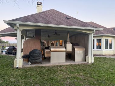 Outdoor kitchen with various grills and a pizza oven under a covered patio with a brown roof.