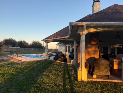 Backyard with a pool, patio, house with dark roof, and lush green grass illuminated by sunlight.