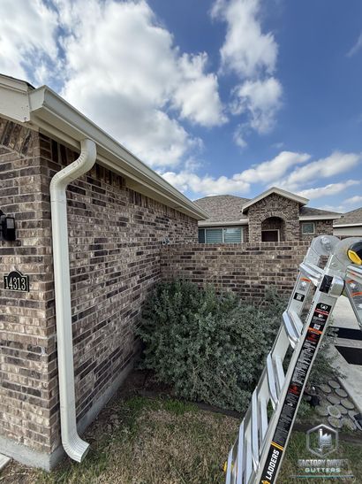 White gutters on brick house, ladder leaned against wall. Blue sky with clouds.