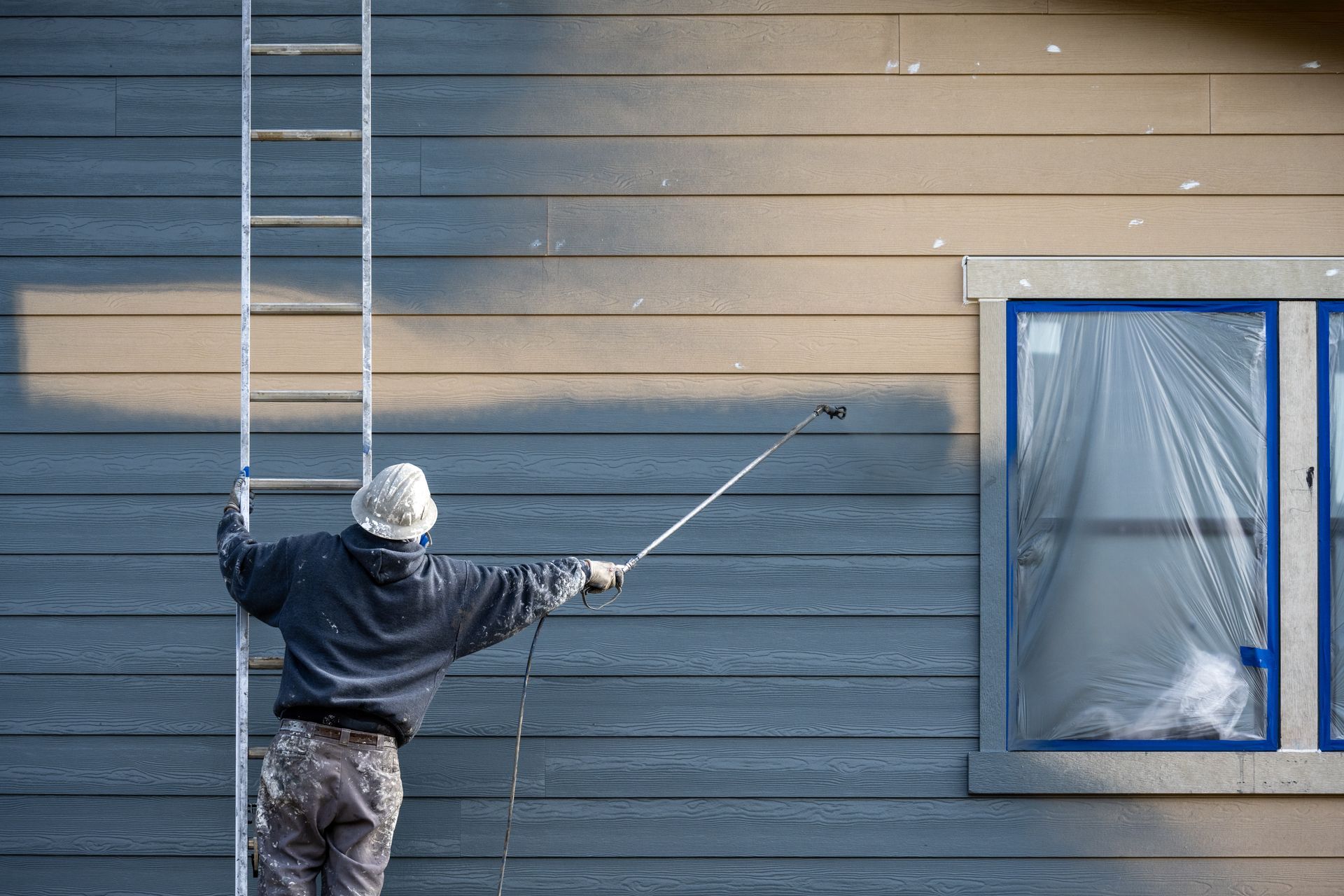 A painter in a hard hat on a ladder with a paint sprayer painting the exterior of a house. A painter in a hard hat on a ladder with a paint sprayer painting the exterior of a house.