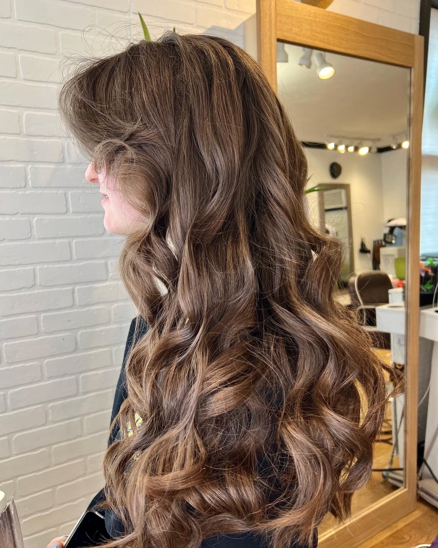 A woman with long brown hair is standing in front of a mirror in a salon.