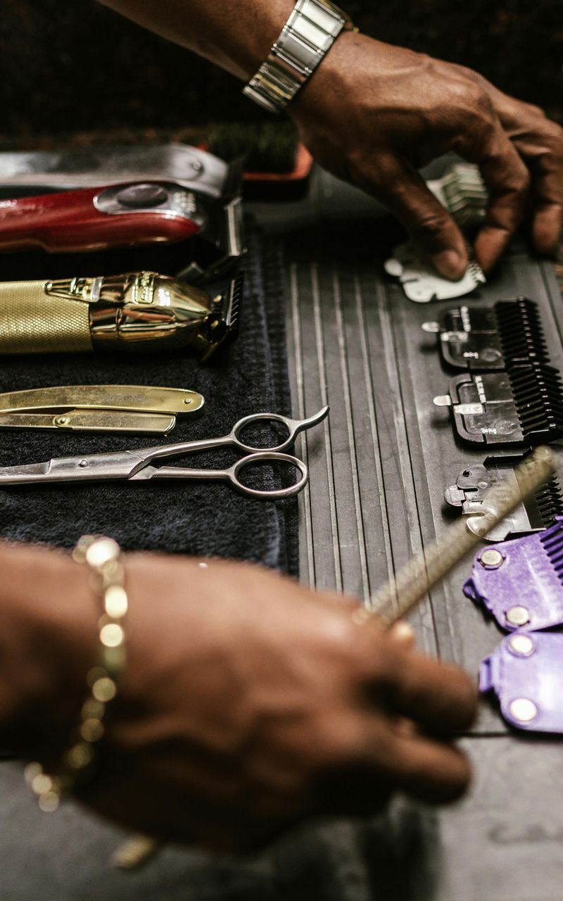 Barber's hands reaching for tools: clippers, scissors, combs, and a brush, displayed on a dark surface.