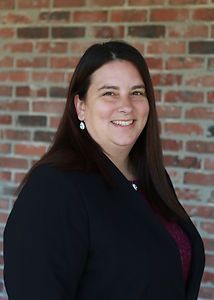 A woman in a black jacket is smiling in front of a brick wall.
