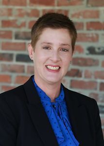 A woman in a blue shirt and black jacket is smiling in front of a brick wall.