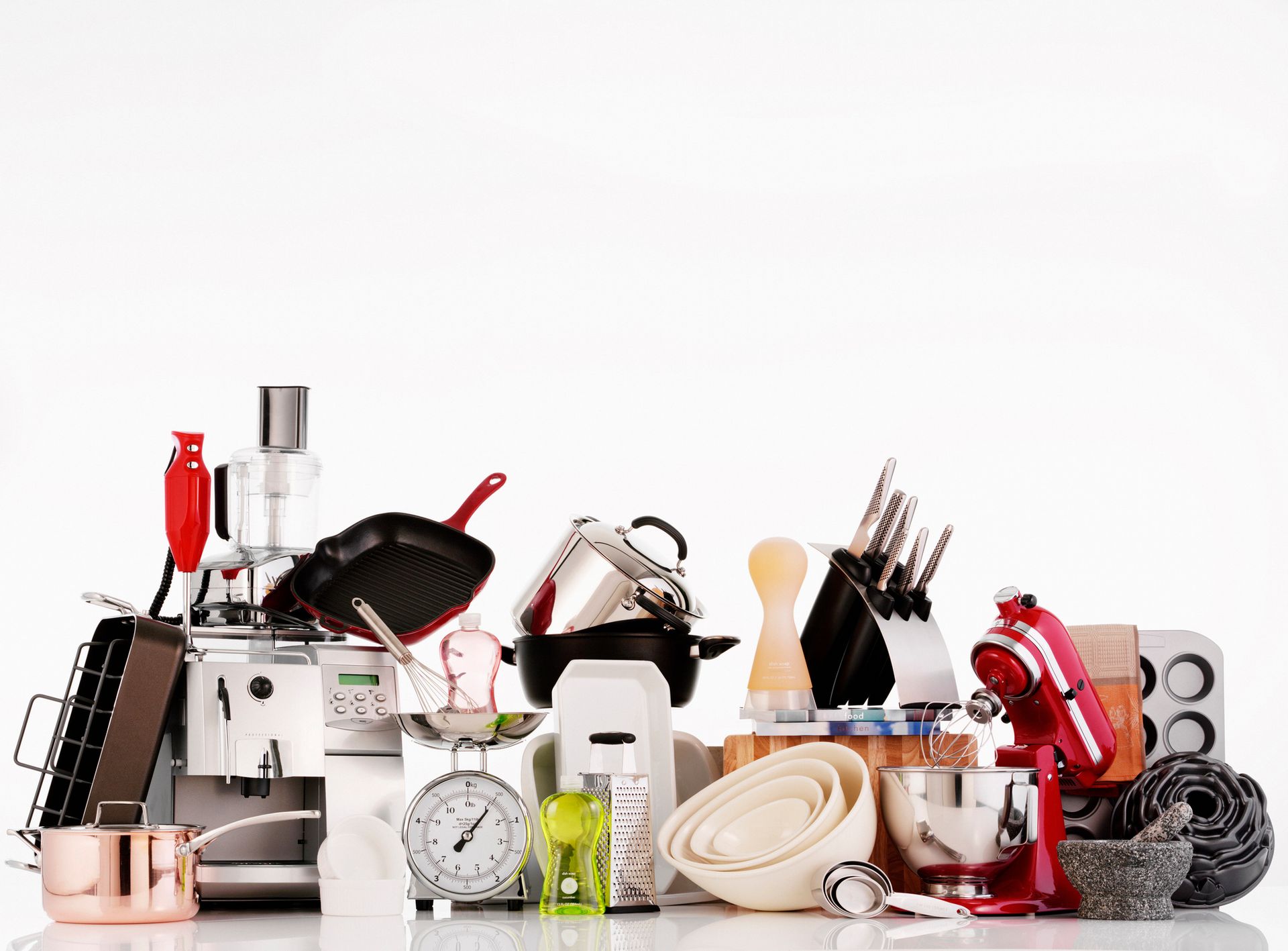 Vari utensili ed elettrodomestici da cucina disposti su una superficie bianca, tra cui un mixer, pentole, padelle e bilance.