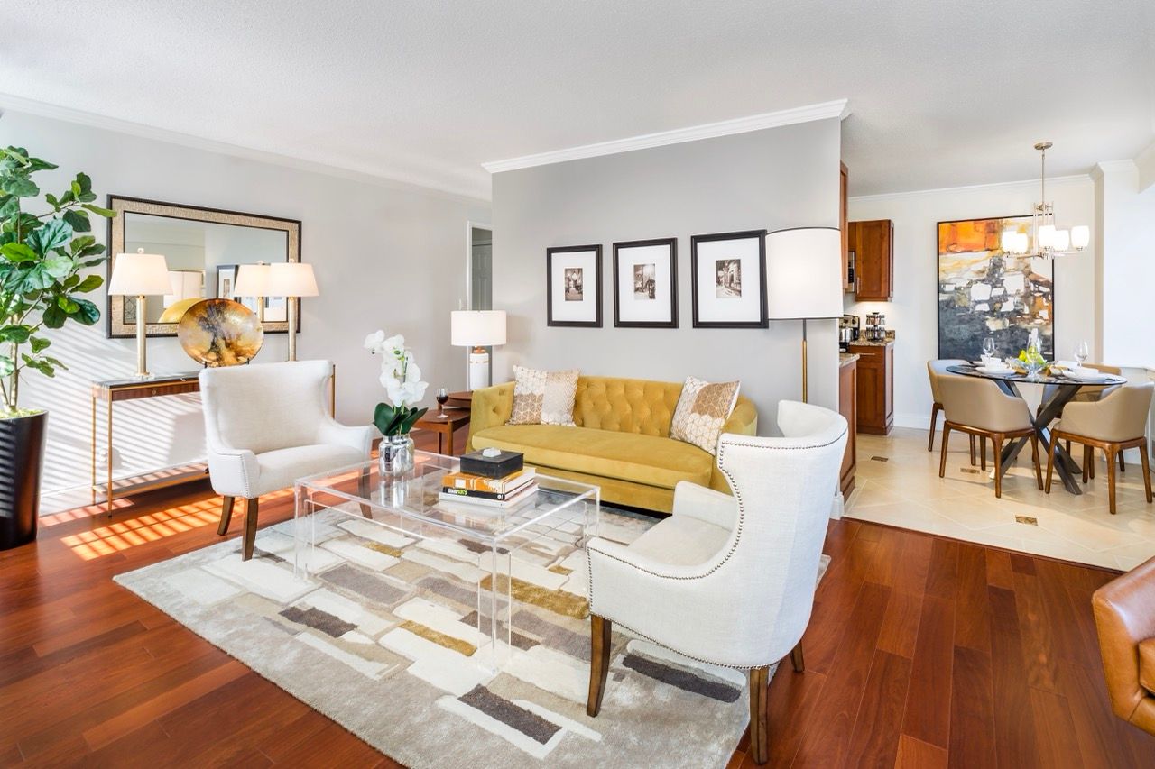 Open-concept living room in a modern apartment with a mustard sofa, white armchairs, glass coffee table, and wall art.
