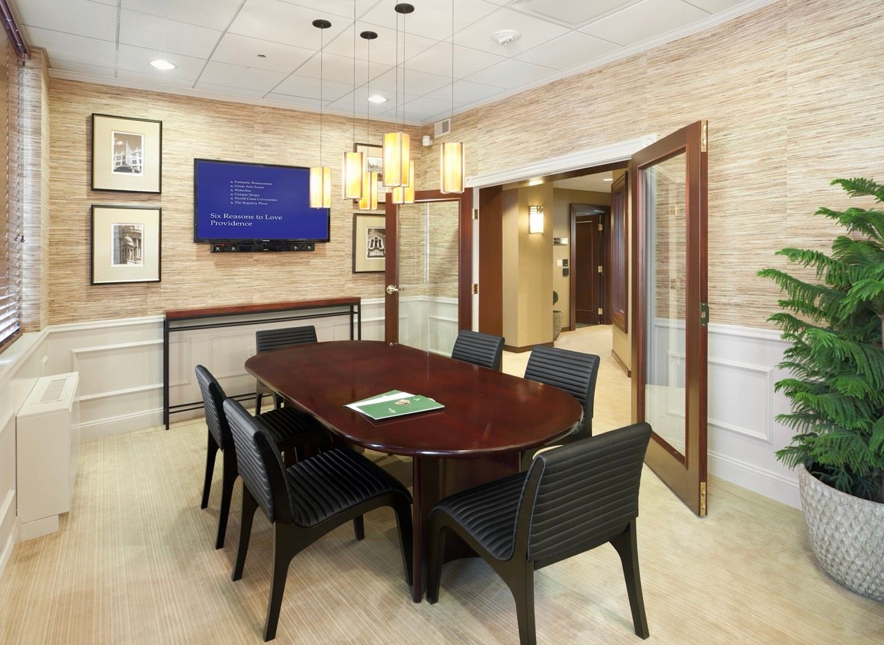 Conference room with an oval dark wood table, black chairs, hanging pendant lights, and a wall-mounted TV.