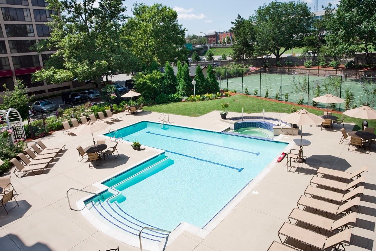 Outdoor pool area with lounge chairs, umbrellas, and a hot tub; adjacent tennis court.