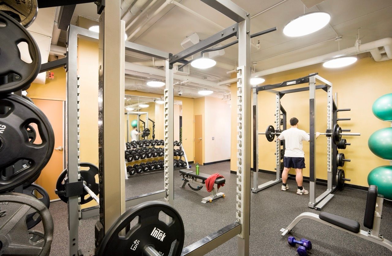 Person standing inside a communal apartment gym near a squat rack and free weights.
