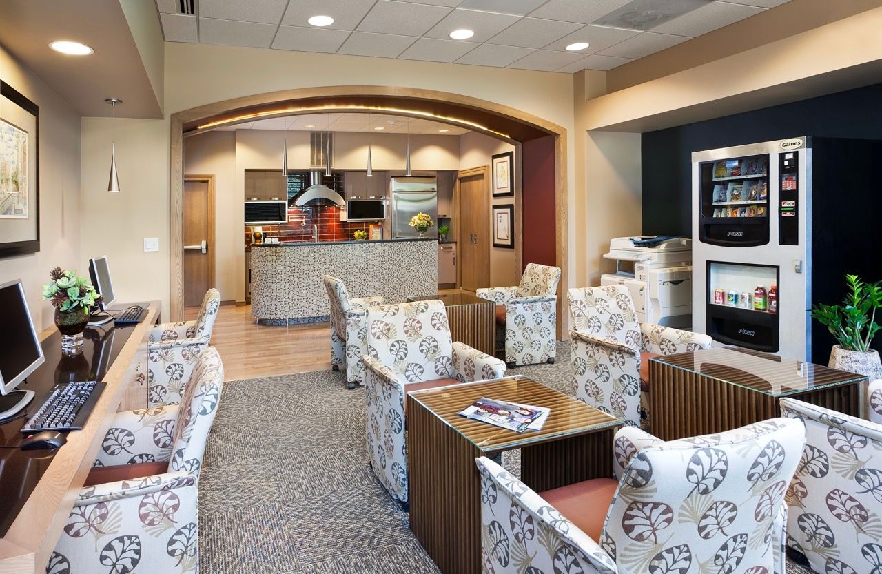 Bright community lounge with seating, a kitchenette, computer desks, and a vending machine.