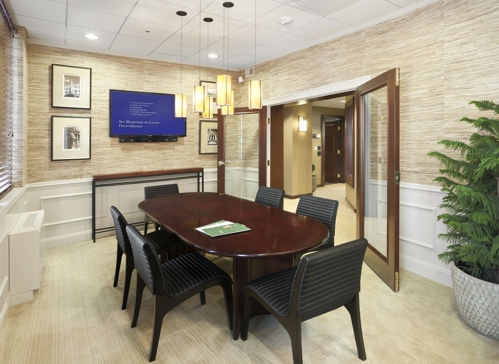 Conference room with an oval dark wood table, black chairs, hanging pendant lights, and a wall-mounted TV.