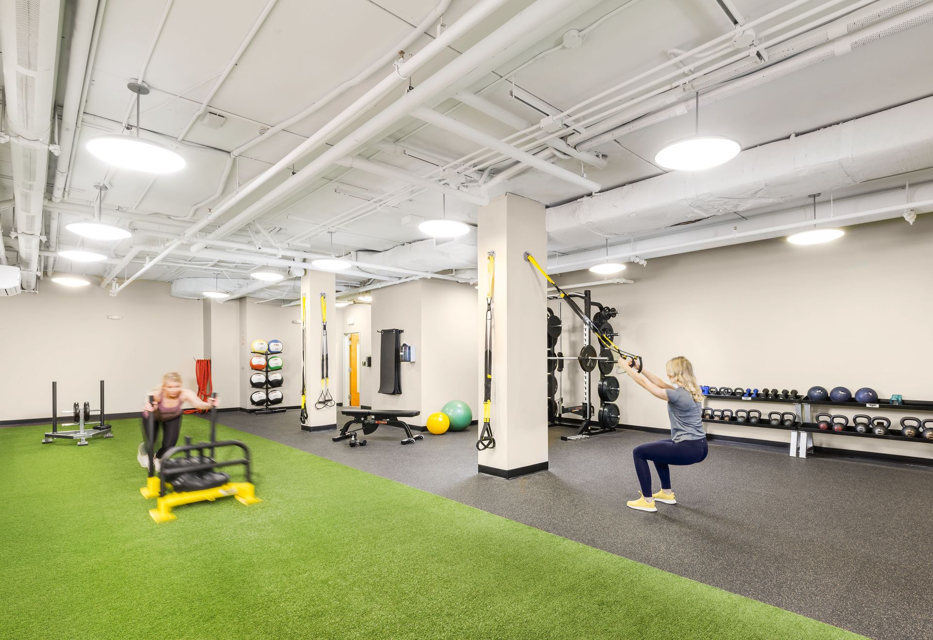 Two people working out in a bright apartment gym with turf, weights, and accessories.
