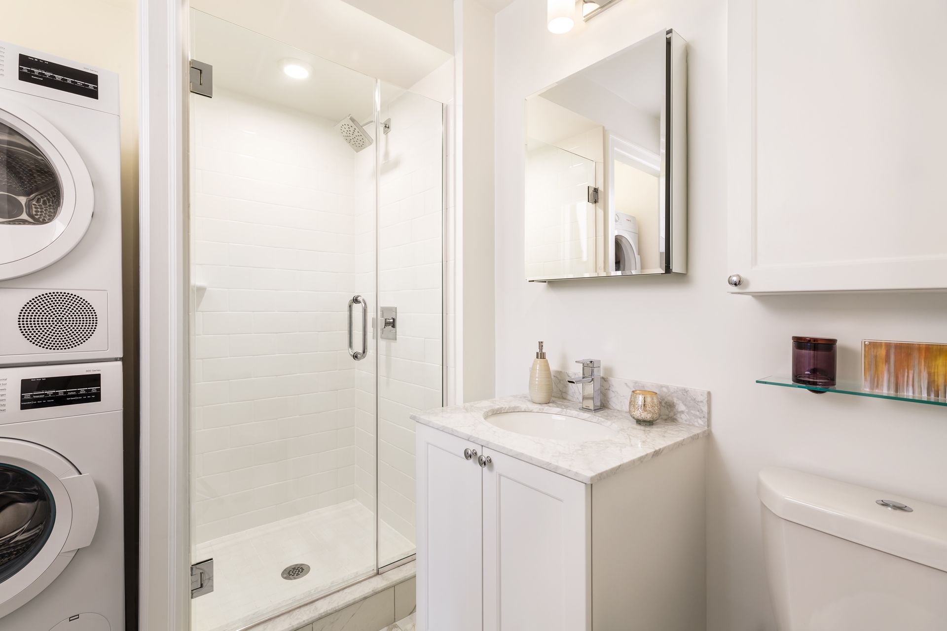 Bright laundry room with stacked washer and dryer, glass shower, vanity, mirror, and toilet.