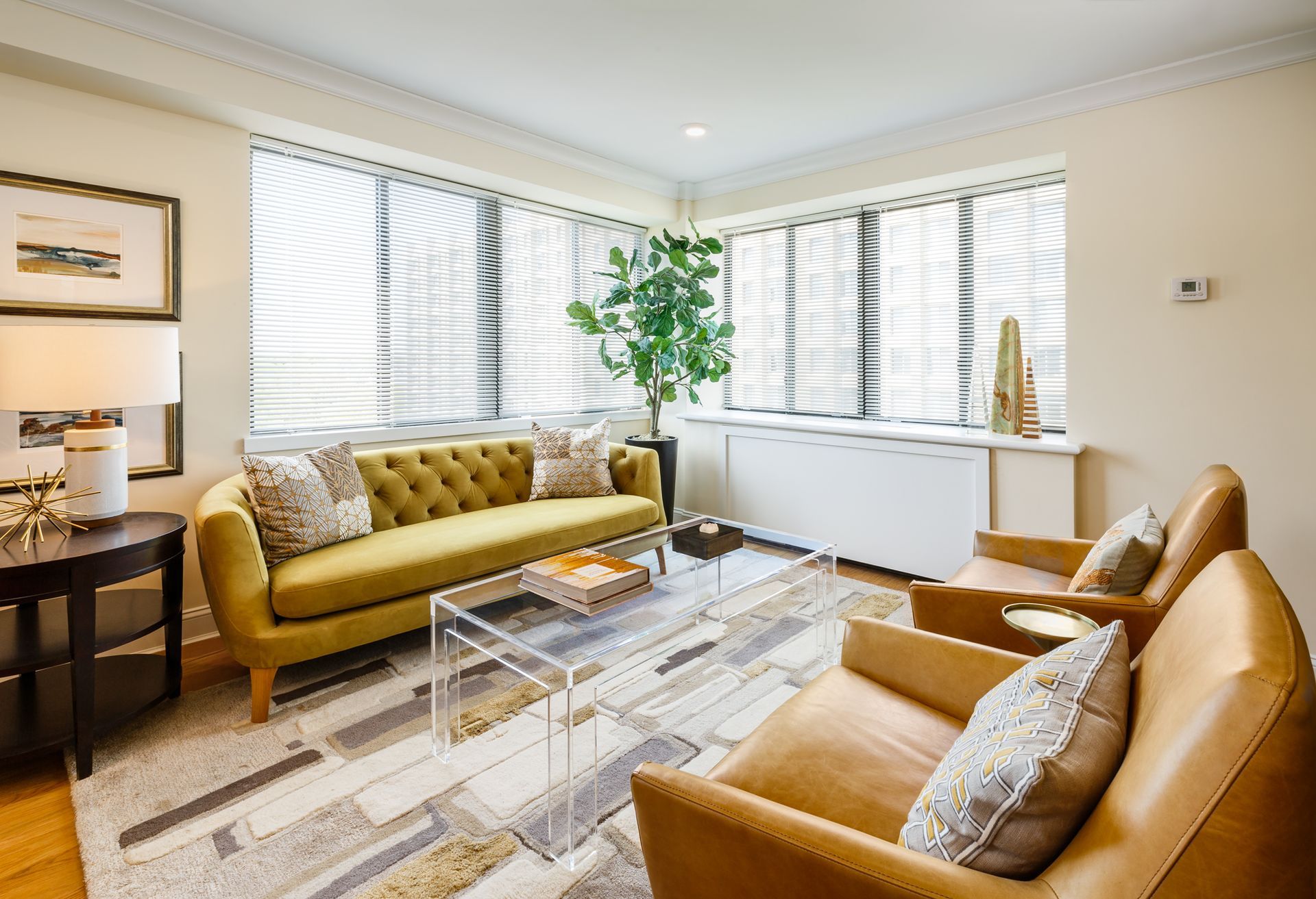 Bright living room with mustard sofa, tan chair, glass coffee table, and large windows with white blinds.