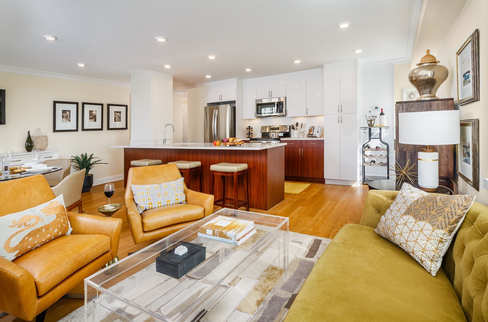 Modern open-plan living room with tan and green sofas, glass coffee table, and a kitchen with island in the background.