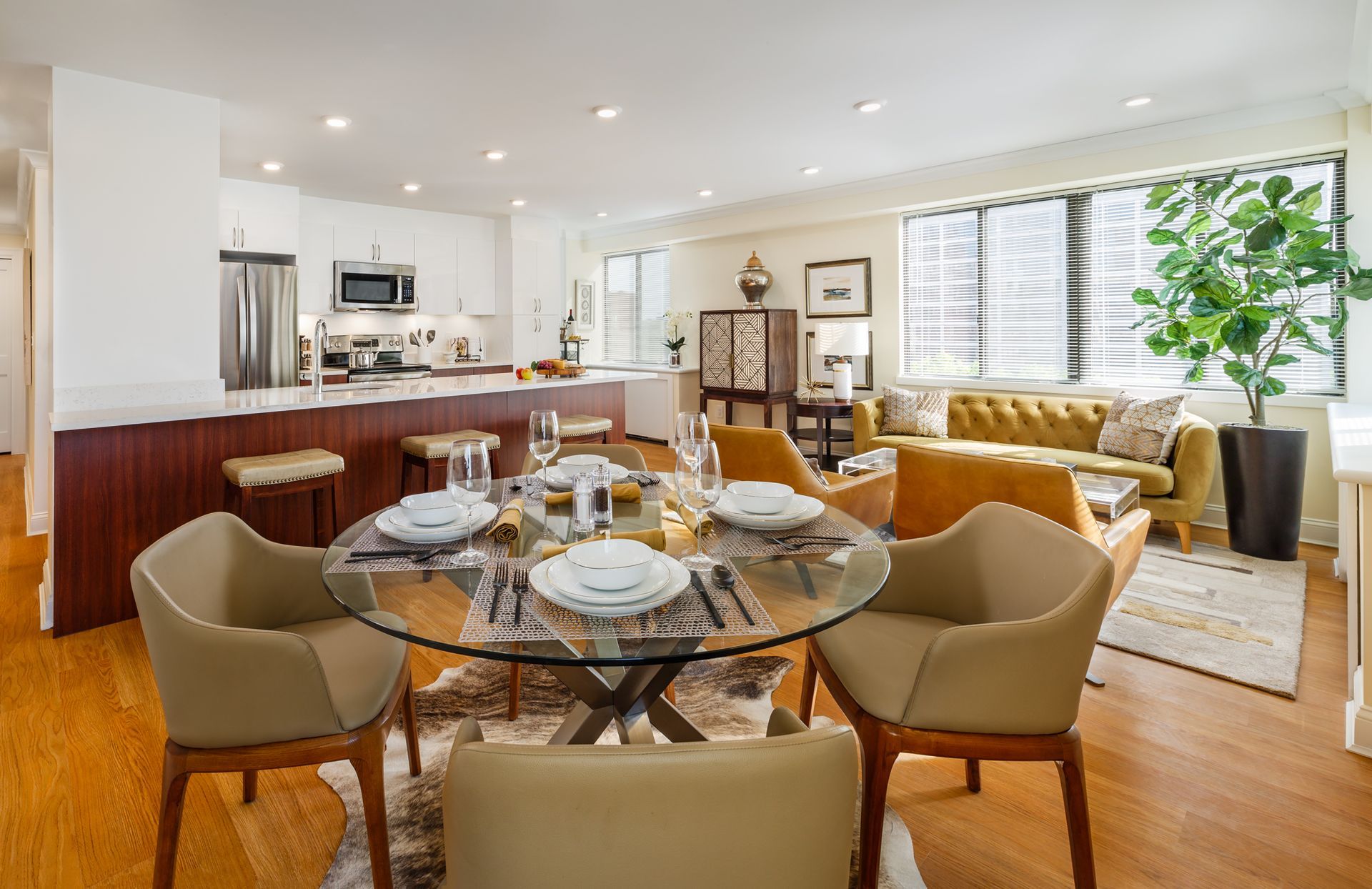 Open-plan living and dining room with glass table, tan chairs, sofa, and kitchen bar seating.