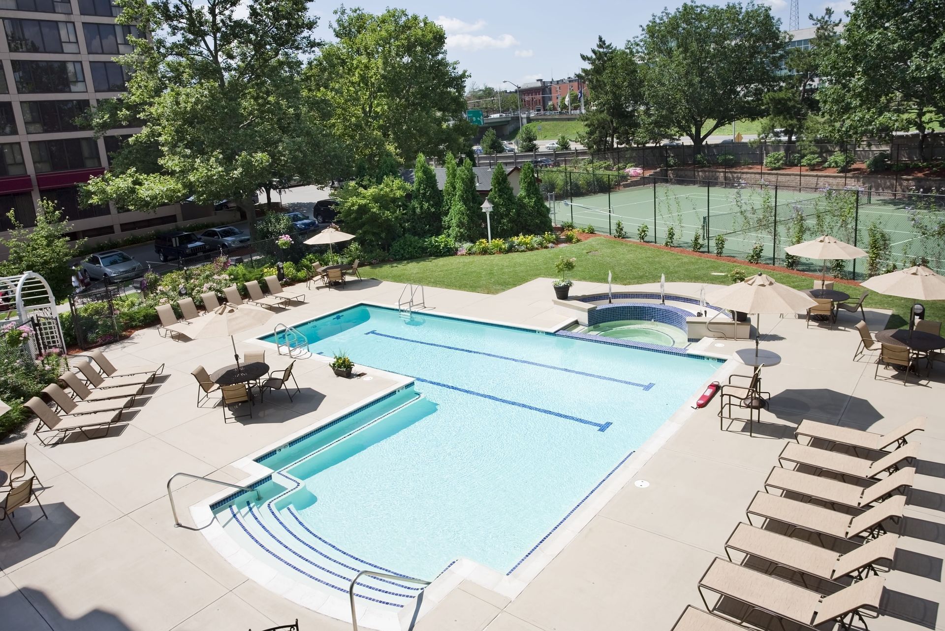 Outdoor pool area with lounge chairs, umbrellas, and a hot tub; adjacent tennis court.