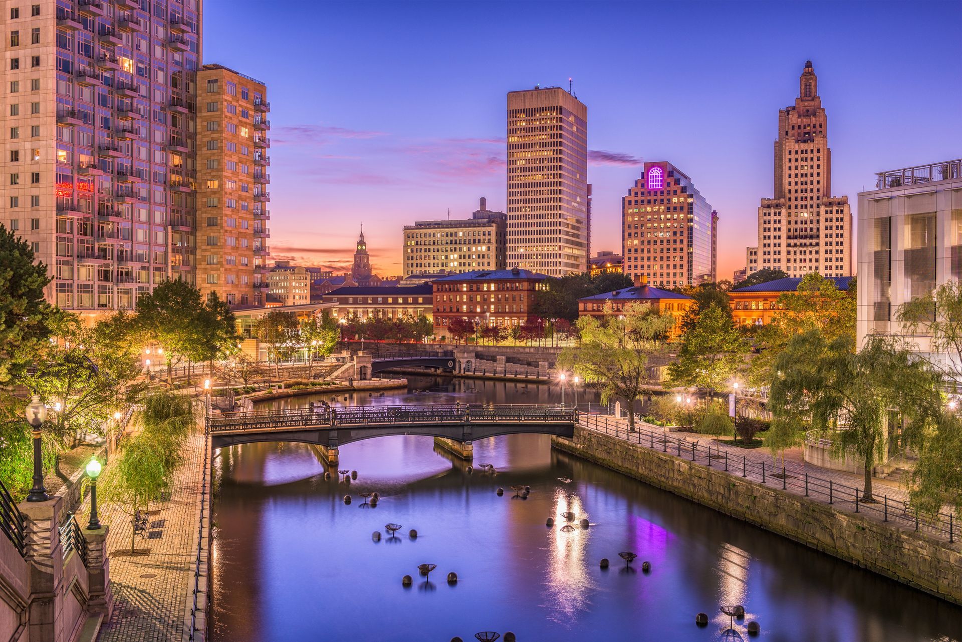 City skyline at dusk with a river, bridges, and colorful lights reflecting on the water