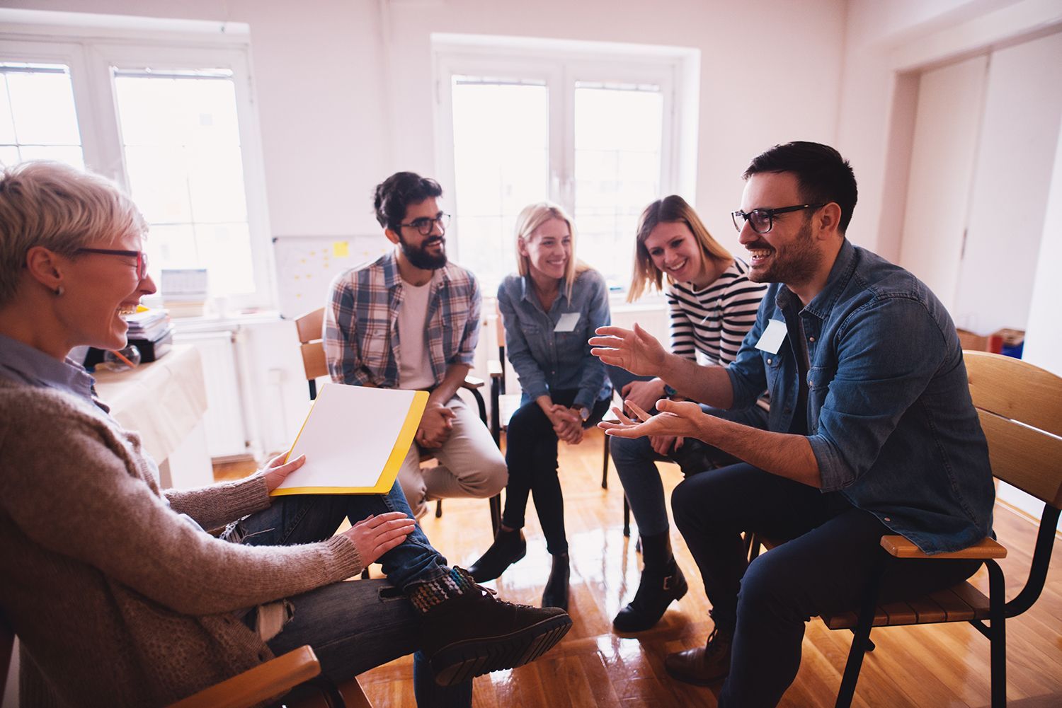 People in a bright room in a group discussion, seated in a circle with notebooks and a laptop.