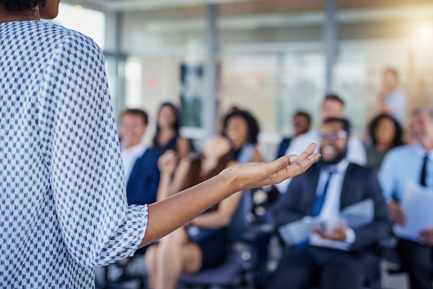 Person speaking to an audience in a bright conference room, gesturing toward seated attendees