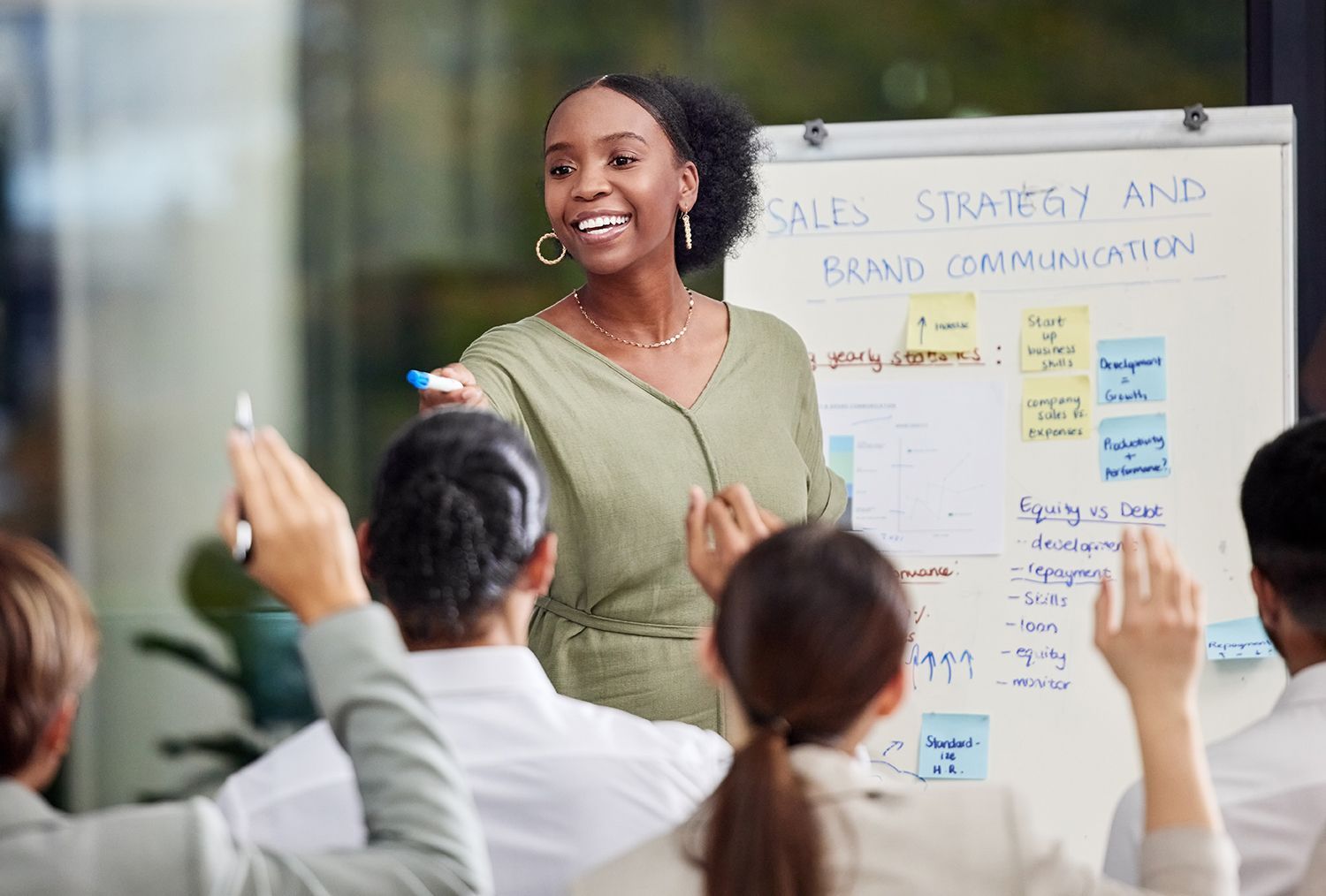 Presenter speaking to a group in a bright classroom, with audience members raising their hands and a whiteboard behind her.