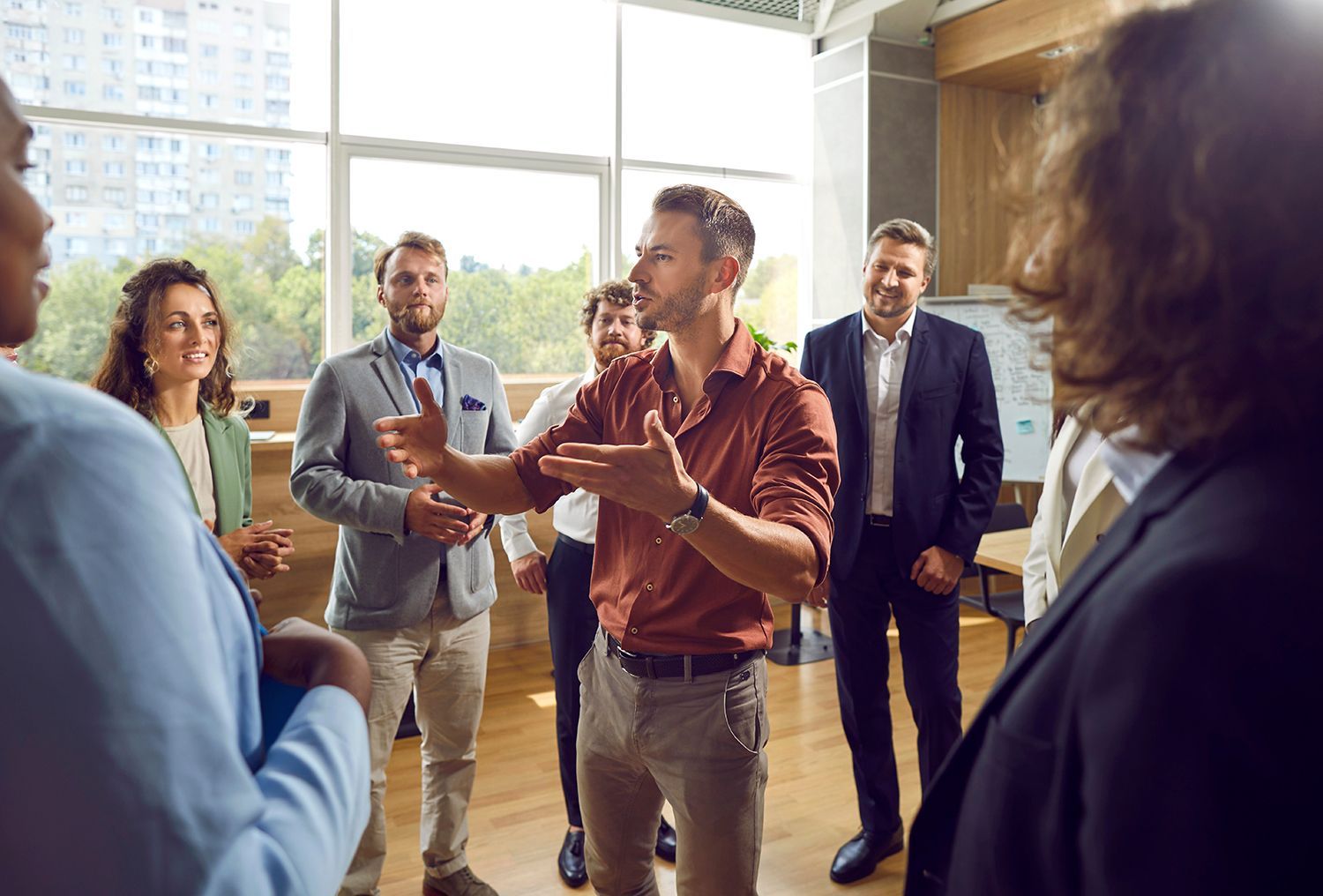 People in a bright office discussing ideas, with one man speaking and gesturing at the center. People in a bright office discussing ideas, with one man speaking and gesturing at the center.