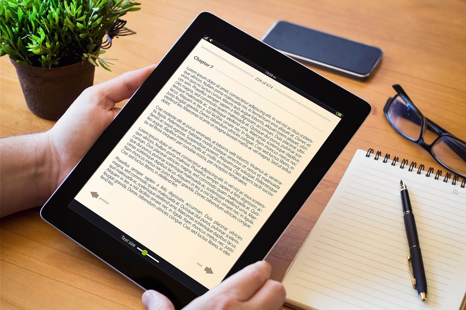Hands holding a tablet displaying text beside a notebook, pen, glasses, phone, and plant on a desk