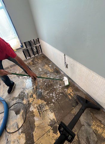 Person scrubbing a wet floor with a mop; vacuum nozzle in foreground; damaged wall.