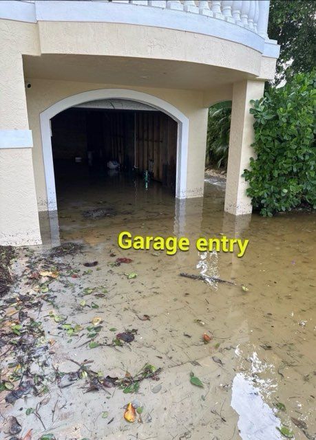 Floodwater surrounds a garage entry; beige structure with white trim. Debris in the murky water.