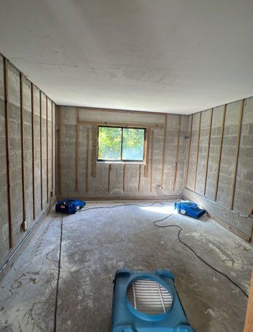 Room under construction; concrete floor, exposed cinder block walls with wood supports, window, and three blue dehumidifiers.