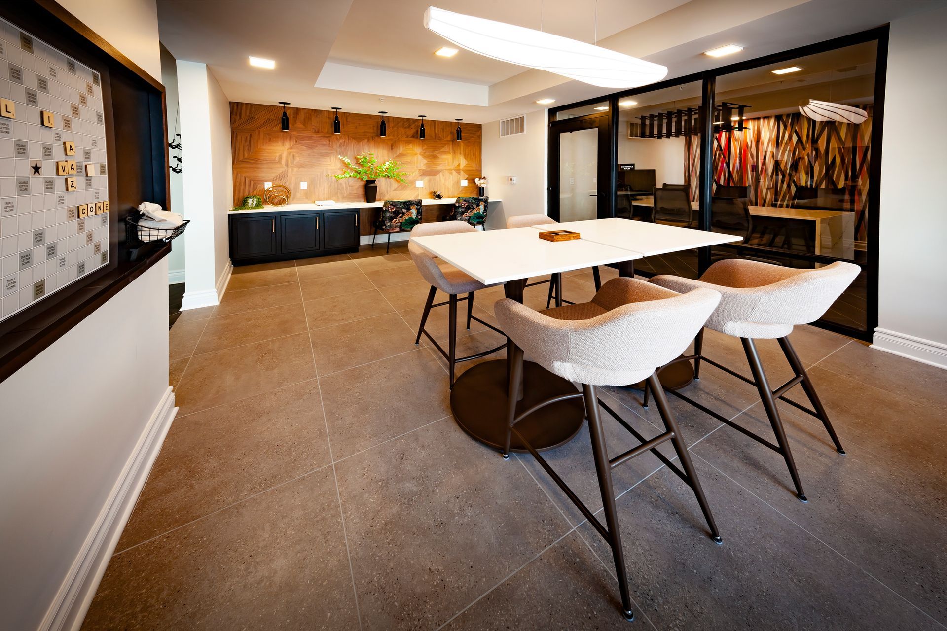 Modern office lounge with a central white table, three high stools, and a back counter area with plants and lighting.