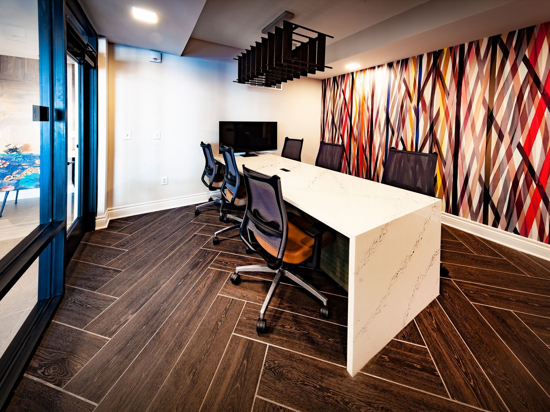 A modern conference room with a white table, office chairs, dark wood-patterned floor, and a colorful abstract wall mural.