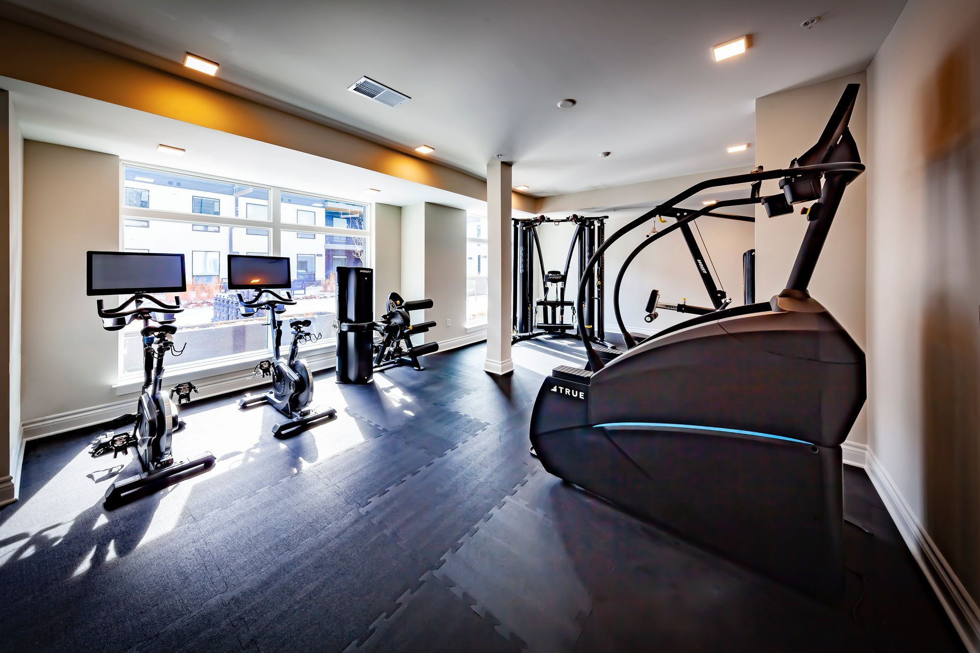 A sunlit fitness room featuring two exercise bikes, a dark elliptical machine, and a weight rack on a rubber floor.
