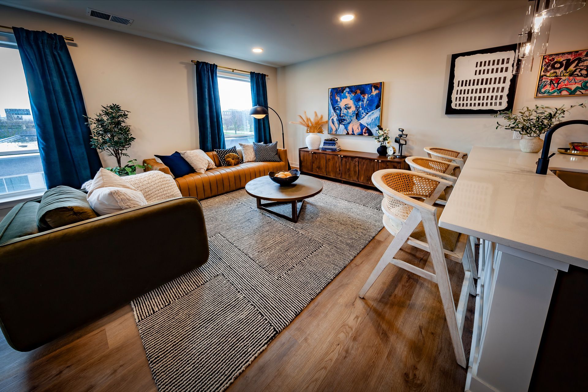 A modern, open-concept living room featuring a patterned rug, a dark green sofa, a tan leather couch, and a kitchen island.