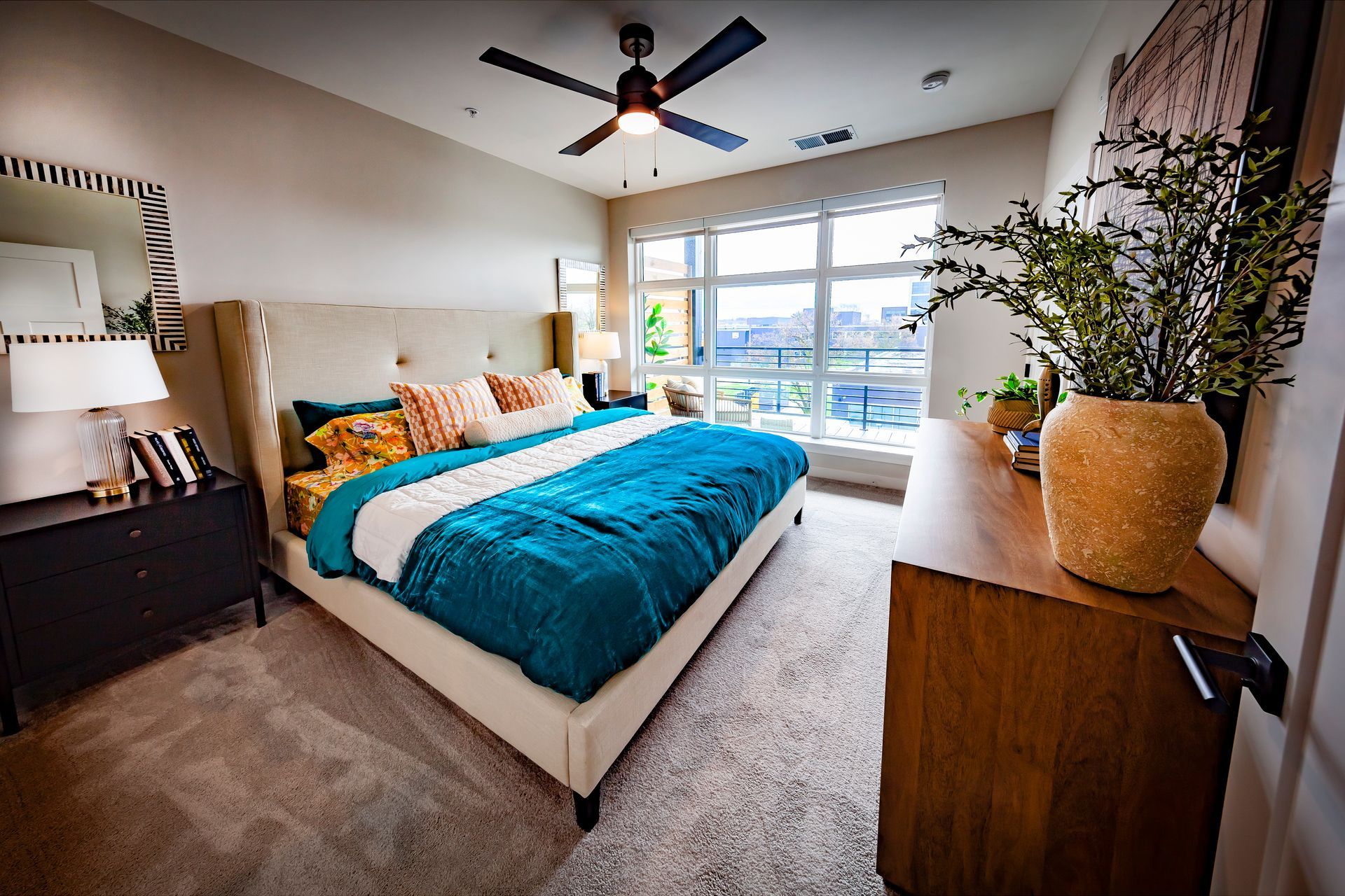 A bedroom with a king-sized bed featuring a bright teal comforter, a wood dresser with a plant, and a large window.