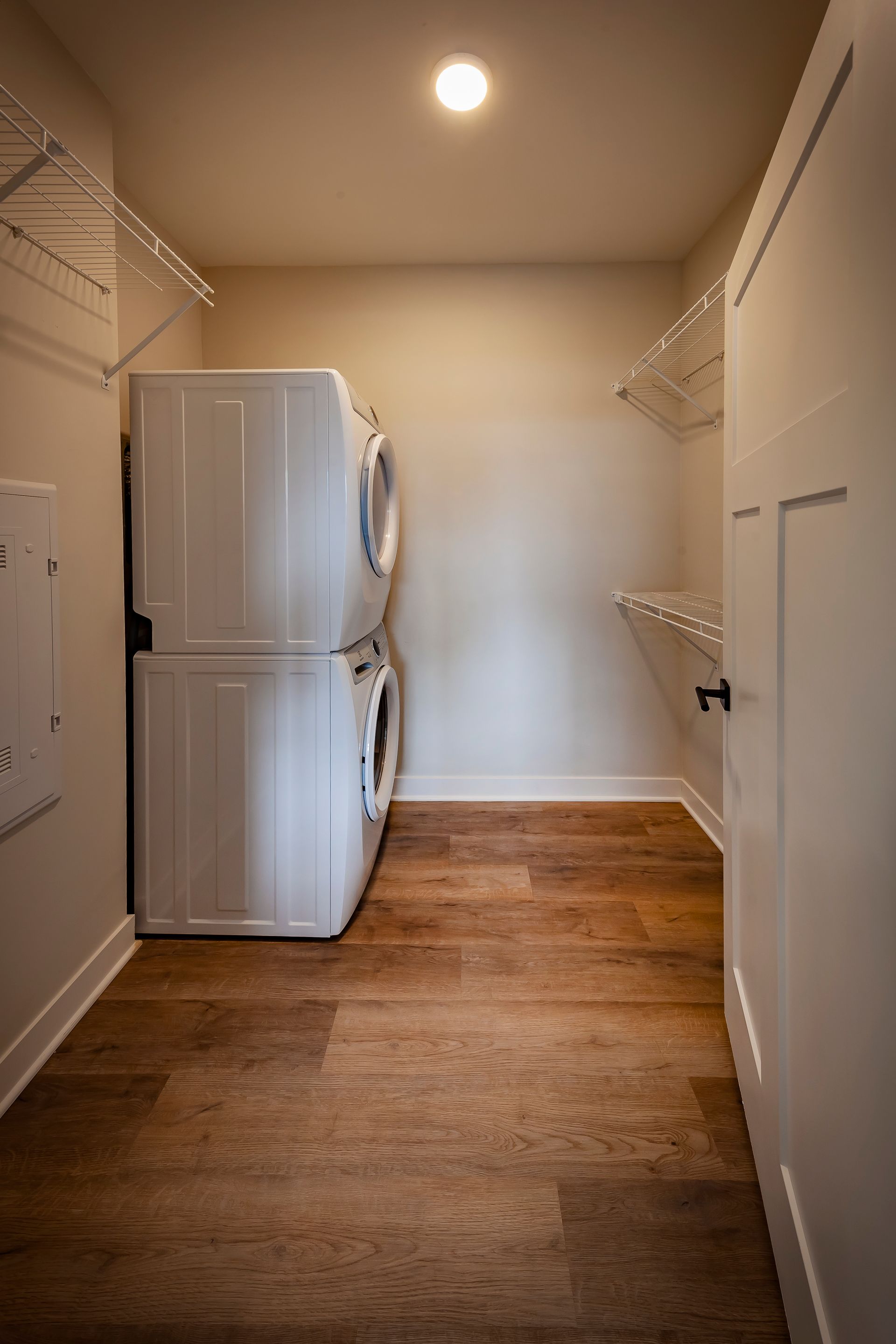 A laundry closet with stacked white appliances on a hardwood floor, flanked by wire shelving units under a ceiling light.