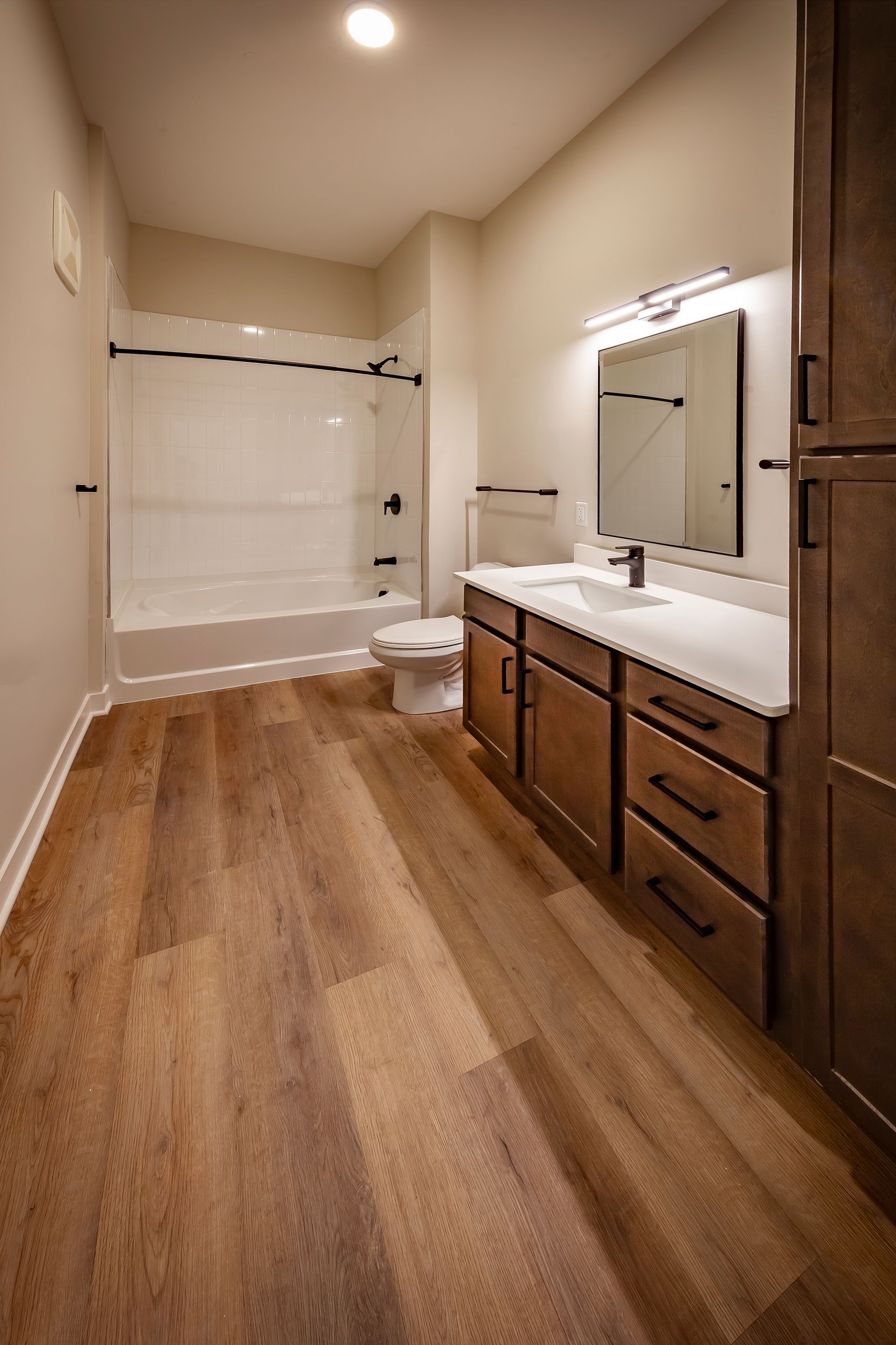 A modern bathroom with wood-look flooring, a white vanity, a mirror, and a shower-tub combo against a cream wall.