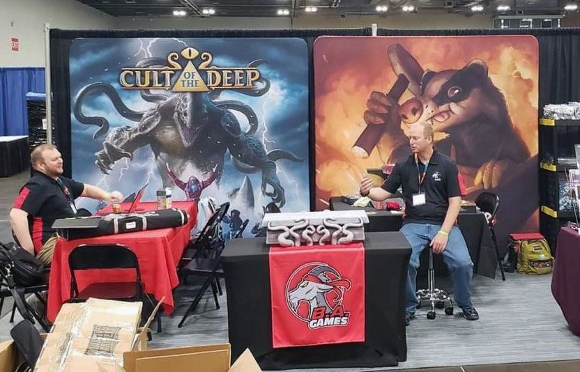 Two men are sitting at a table in front of a sign that says cult of the deep.