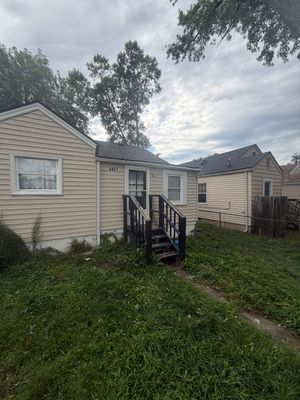Small beige house with black front stairs and overgrown grass in the yard