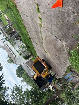 Yellow utility truck beside orange cone on a cracked driveway near a house and trees
