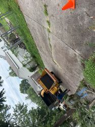 Yellow dump truck on a cracked concrete lot near grass and a chain-link fence, with an orange cone nearby.