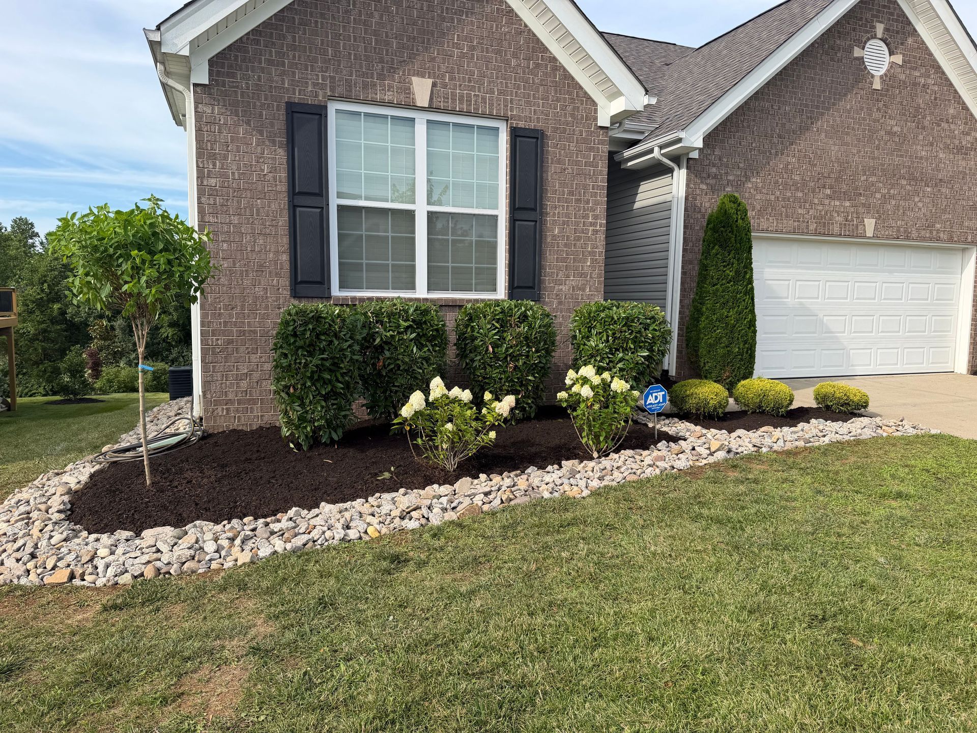 Suburban brick house front yard with mulch bed, shrubs, and white garage door