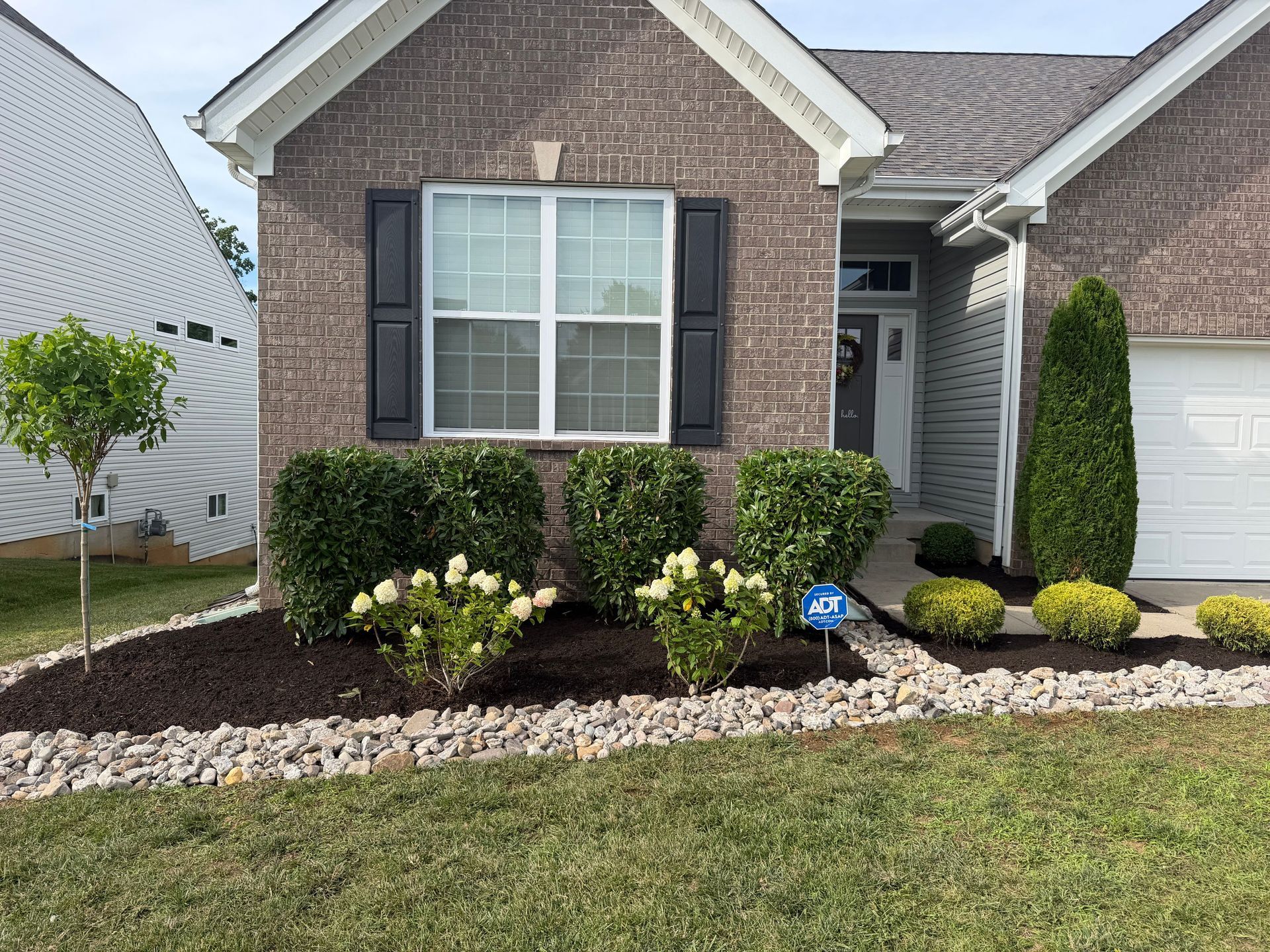 Brick suburban house front with black shutters, white garage, and trimmed shrubs along the walkway