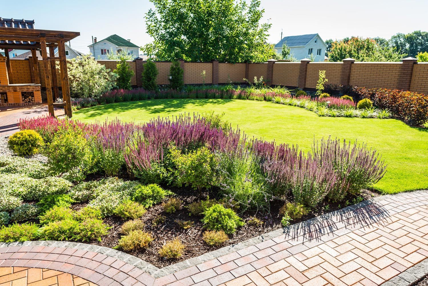 Landscaped backyard garden with purple flowers, green lawn, brick patio, and wooden pergola under sunny skies