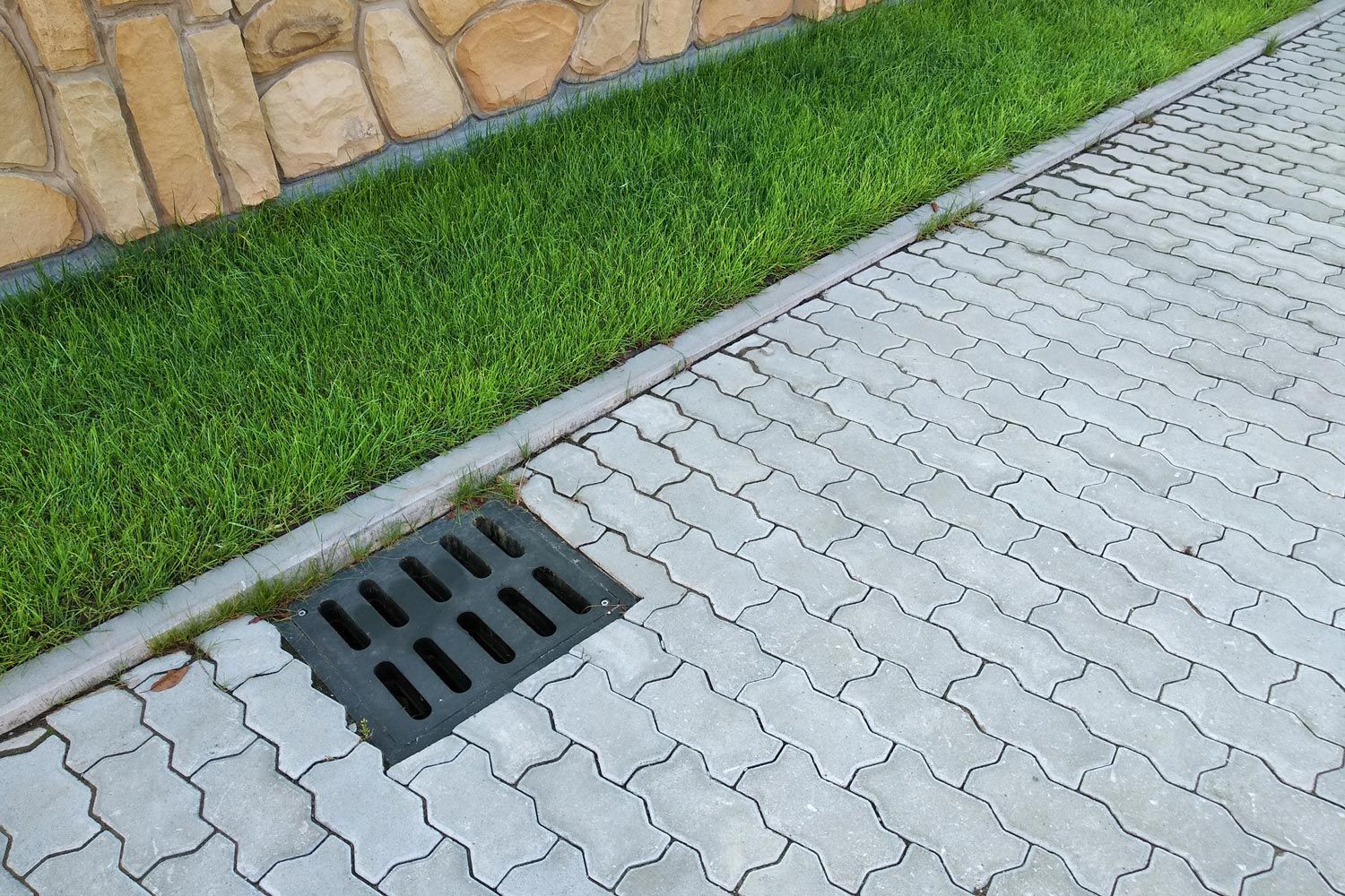 Storm drain grate beside a grassy strip and tan stone wall along a paved walkway.