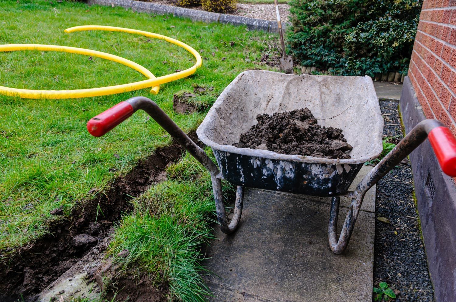 Wheelbarrow full of soil on a concrete path beside a grassy yard with a yellow hose and brick wall.