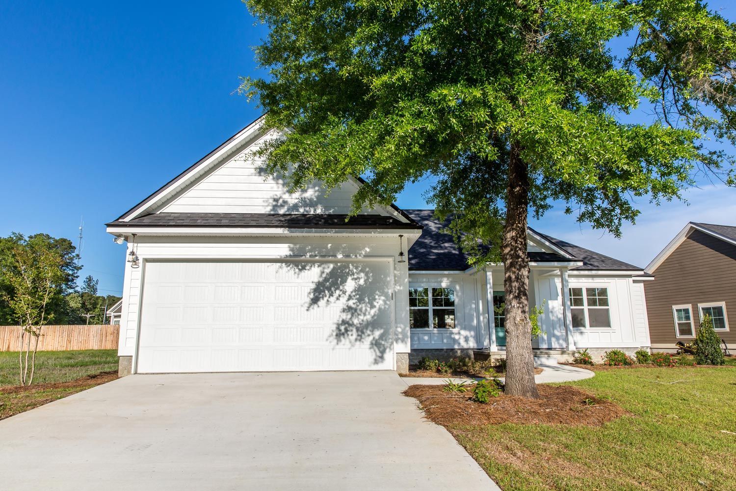 Suburban house with white garage, large tree, and driveway under a clear blue sky Suburban house with white garage, large tree, and driveway under a clear blue sky